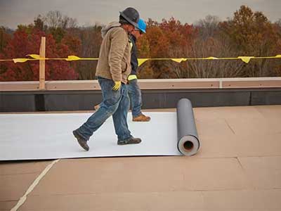 Two workers on a flat commercial roof rolling out a white TPO/PVC single-ply sheet